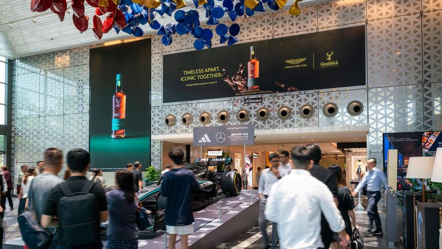 Two JCDecaux digital screens over the entrance to Raffles City, running a whisky campaign above a Formula One car on display, with shoppers walking past.