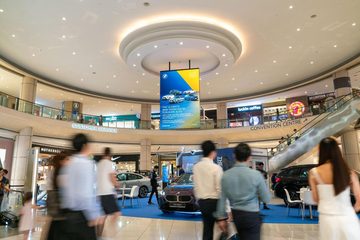 A digital screen hanging in the atrium of Suntec City, showing a car showcase advert above the cars on display, with shoppers walking past.