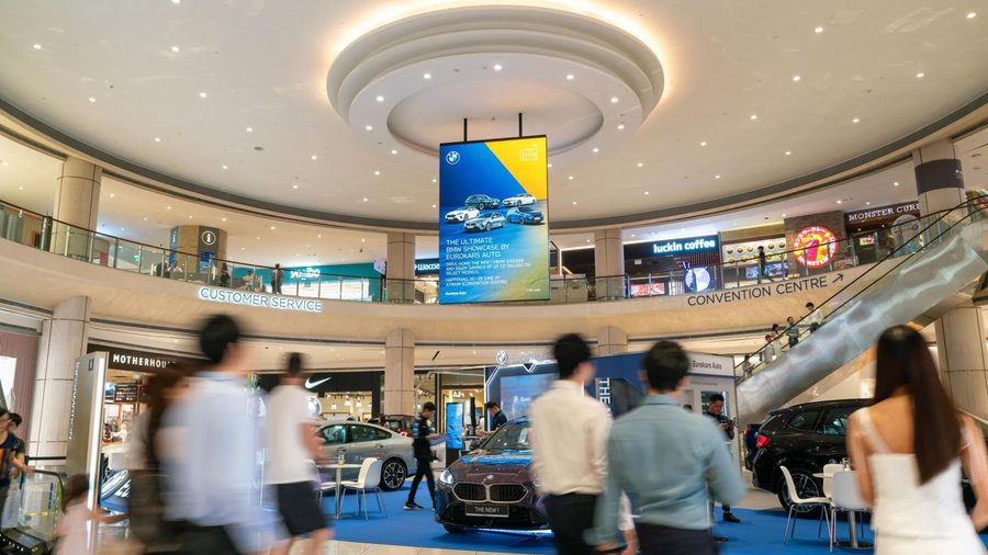 A digital screen hanging in the atrium of Suntec City, showing a car showcase advert above the cars on display, with shoppers walking past.