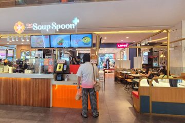 A The Soup Spoon+ counter in a shopping mall: the flower mark and the name above three menu screens, a customer ordering at the counter, and diners at the tables beside it.
