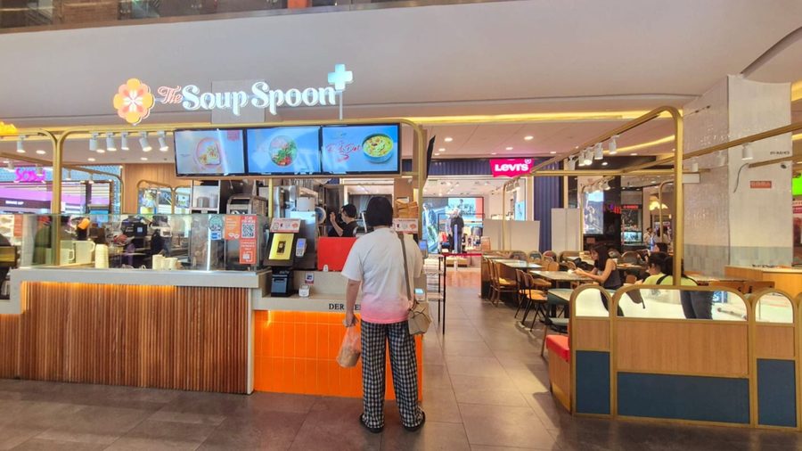 A The Soup Spoon+ counter in a shopping mall: the flower mark and the name above three menu screens, a customer ordering at the counter, and diners at the tables beside it.