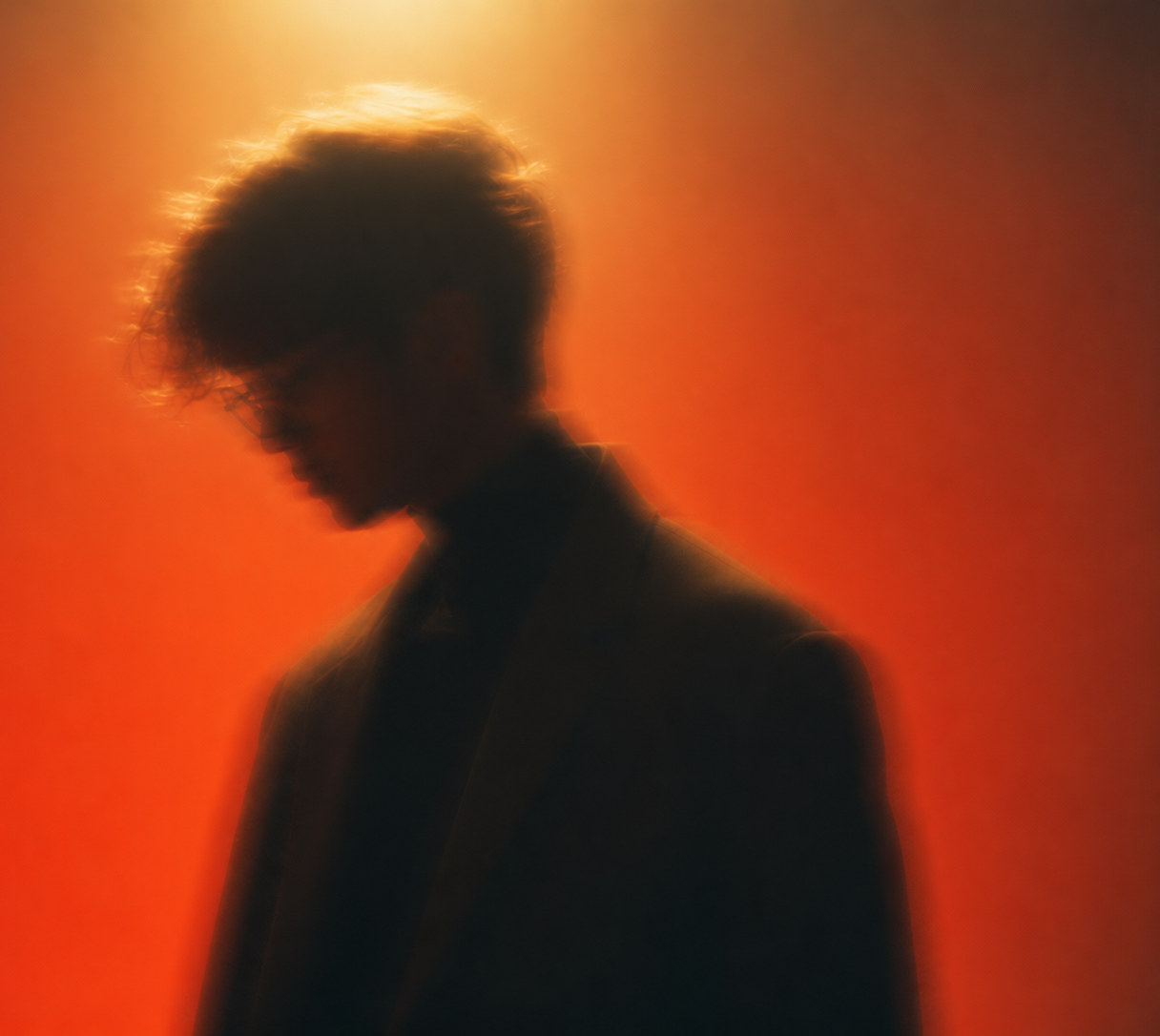 Tham Yu Le in profile, head lowered, lit from behind against a deep orange field.
